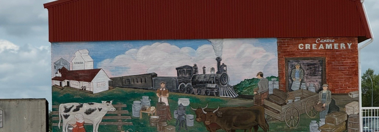 Colourful mural on a red-roofed building showing a historical creamery scene with cows, train, and workers, labelled “Canora Creamery.”