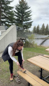 Ella tying her hiking boots on a picnic table beside a suspension bridge and river before starting a trail walk.