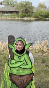 Ella posing behind a painted green trail troll cutout beside the water near a bridge in Wolseley, Saskatchewan.