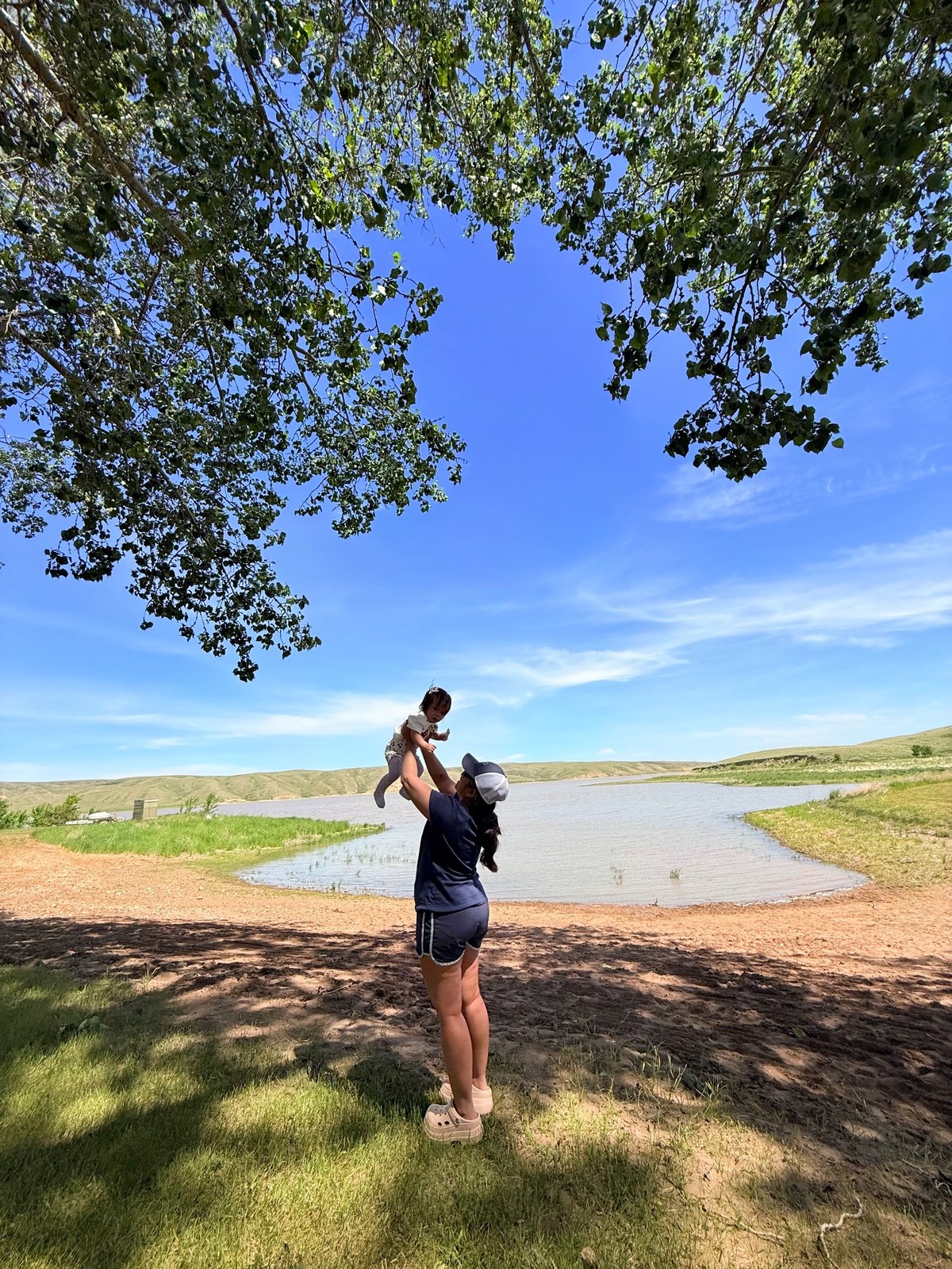 An adult lifting a young child beside a prairie lake on a sunny Saskatchewan day.