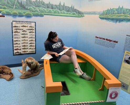 Woman reading a brochure while sitting in a model boat at the Duck Mountain Provincial Park visitor center with educational displays behind her.