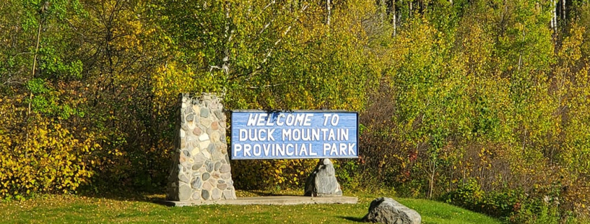 Blue and white "Welcome to Duck Mountain Provincial Park" sign with a stone pillar base, surrounded by green grass and yellow autumn trees.