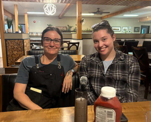 April, co-owner of Duck Mountain Lodge, sitting with Ella inside the lodge dining area with ketchup, salt and pepper on the table in front.