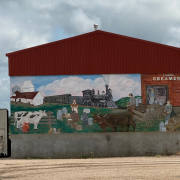 Colourful mural on a red-roofed building showing a historical creamery scene with cows, train, and workers, labelled “Canora Creamery.”