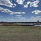 Wide view overlooking Wascana Habitat Conservation Area from Douglas Park hill, with water, prairie grass, and a bright blue Saskatchewan sky filled with clouds.